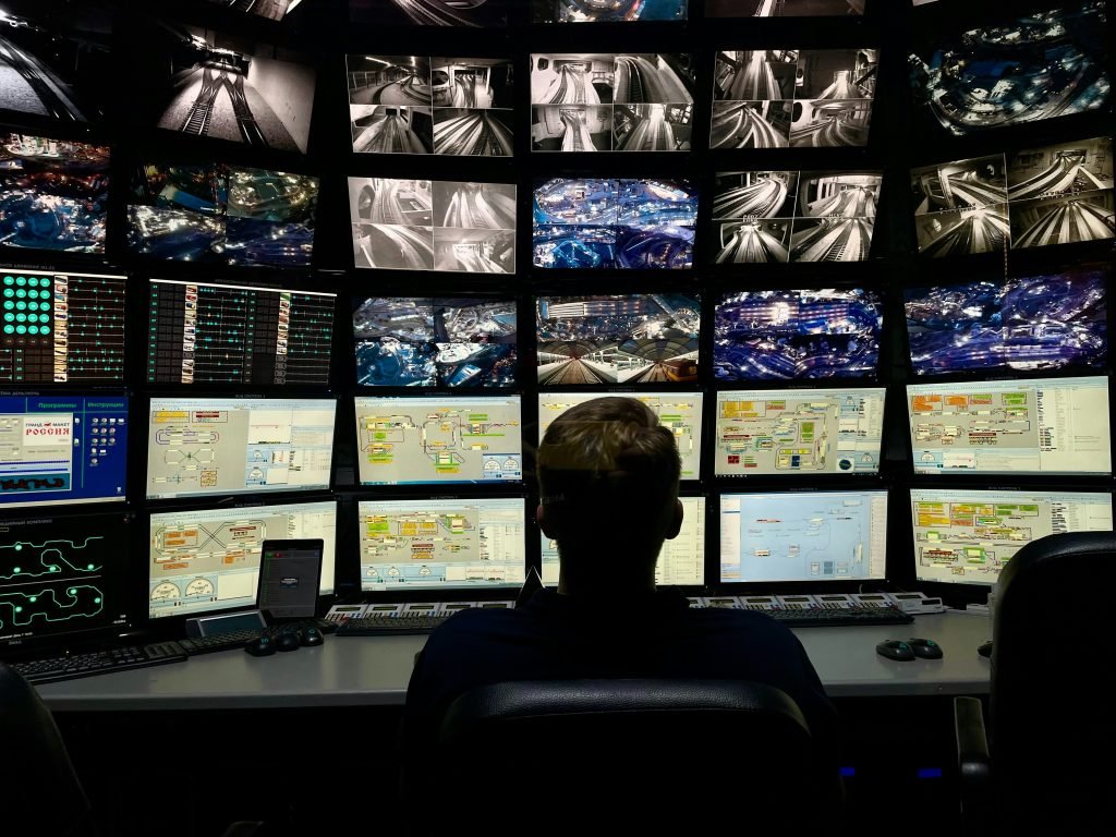 Operator seated at a wall of dozens of monitors showing live network maps, camera feeds, and system dashboards in an enterprise command center