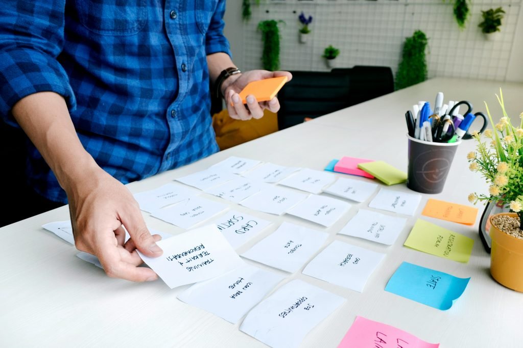 A hand arranges handwritten sticky notes on a desk during a project planning card-sorting exercise, with pens and a small plant nearby